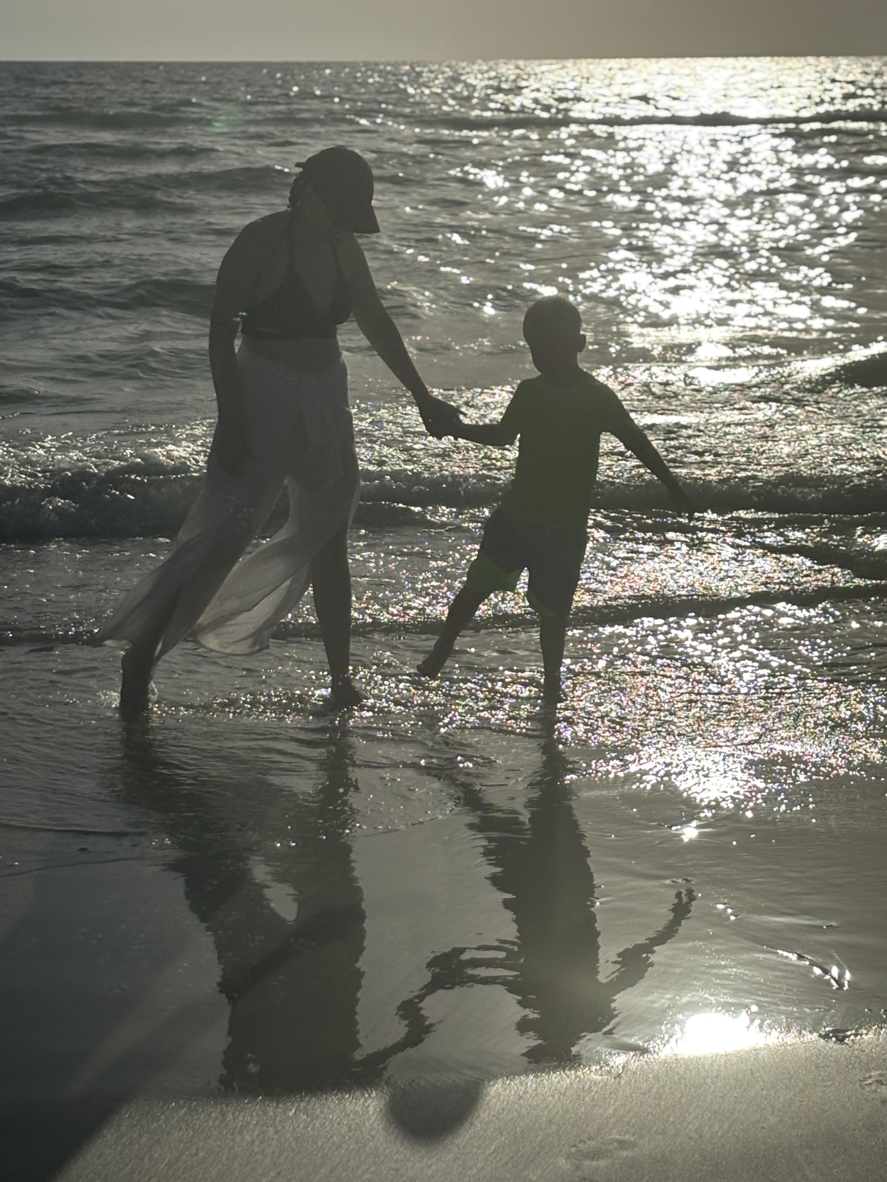 Silhouette of a mother and child holding hands at the water's edge at sunset